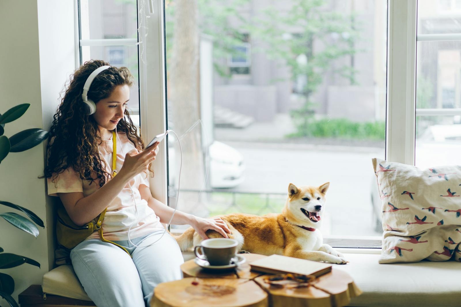 Woman listening to music on her phone while sitting by the window with her dog in a cozy home setting.