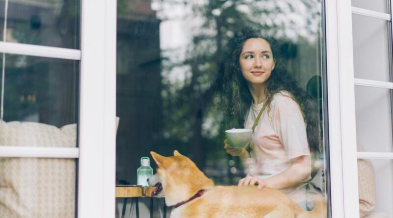 Relaxed woman enjoying a warm beverage with a Shiba Inu dog by a cozy window view.