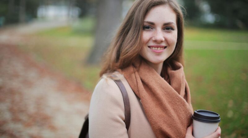 Portrait of a smiling woman holding a coffee cup in a serene autumn park setting.